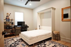 Ein Schlafzimmer mit einem Bett, einem Schreibtisch, einem Stuhl, einem kleinen Hocker, einem Deckenventilator und einem Fenster.