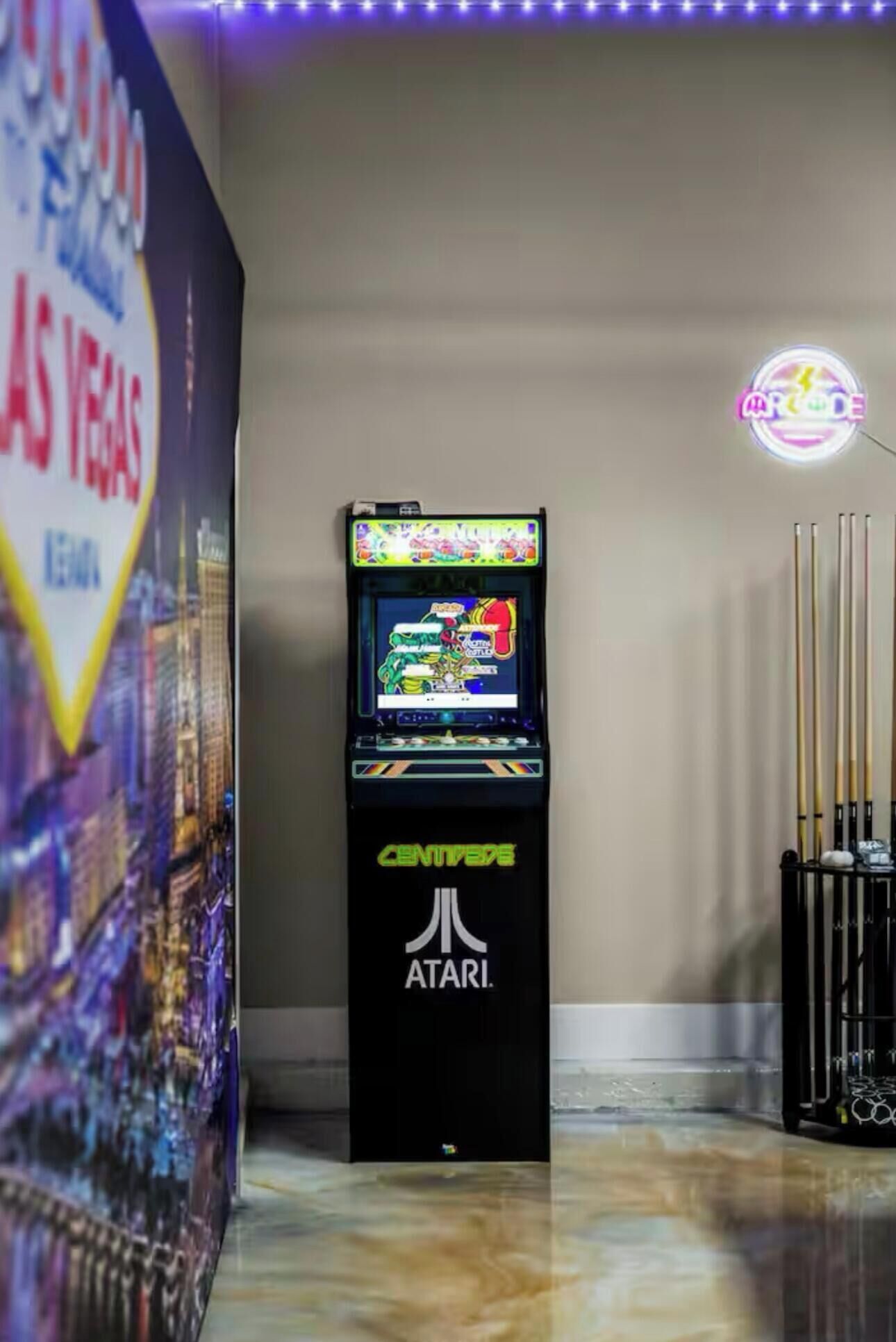 Máquina de videojuegos con una pantalla colorida, ubicada en una sala con mesa de billar y un letrero de neón.
