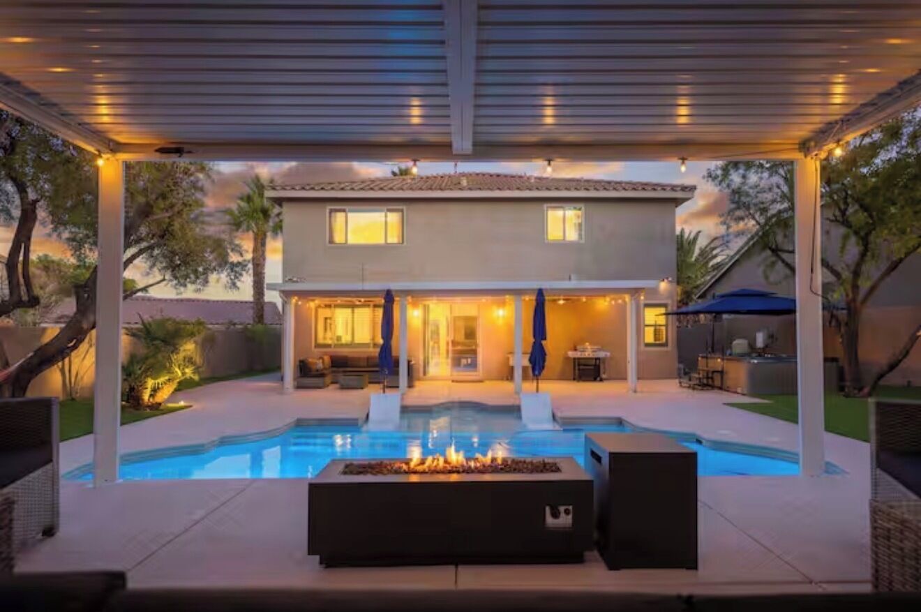 A covered outdoor swimming pool area with a fire feature, a patio with seating, and a house in the background.