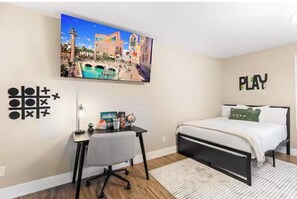 Ein Schlafzimmer mit einem Bett, einem Schreibtisch und einem an der Wand montierten Fernseher, der eine Stadtansicht zeigt.