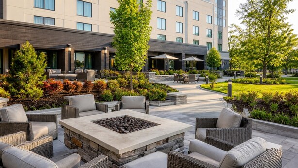 A modern outdoor seating area with wicker furniture and a stone fire pit, surrounded by landscaped greenery and a building in the background. - NovaStar Test Hotel od0mjx (Springfield)