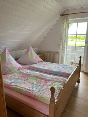 Una cama individual con estructura de madera, colchón rosa y colcha de cuadros. Se ve vegetación a través de la ventana.