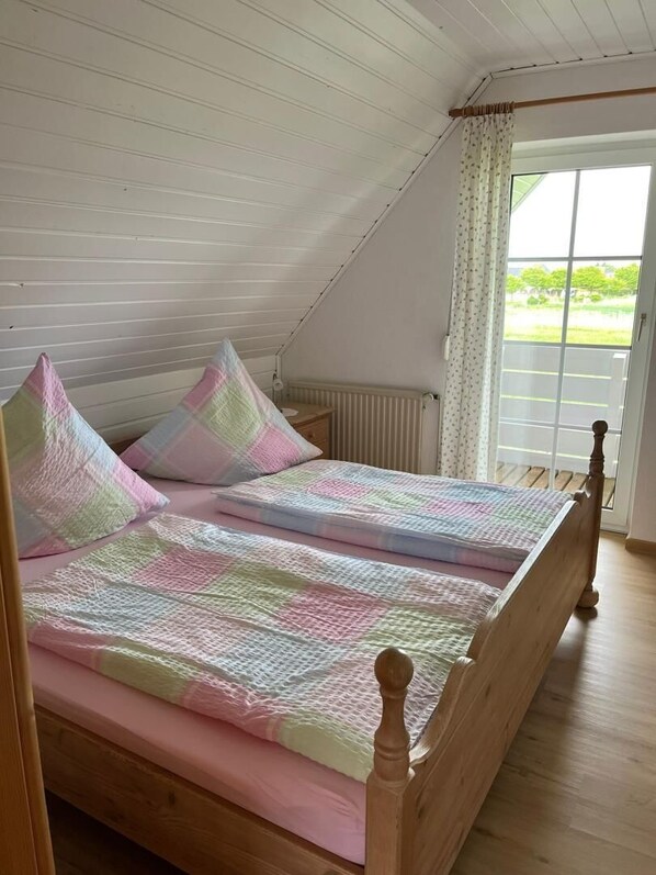 Un lit simple avec un cadre en bois, un matelas rose et une couette à motifs à carreaux. Une fenêtre donne sur un paysage verdoyant.