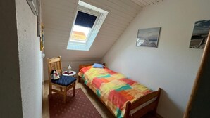 Ein kleines Zimmer mit einem Einzelbett, einem Stuhl, einem Tisch, einem Fenster mit Blick und einer Wand mit zwei Bildern.