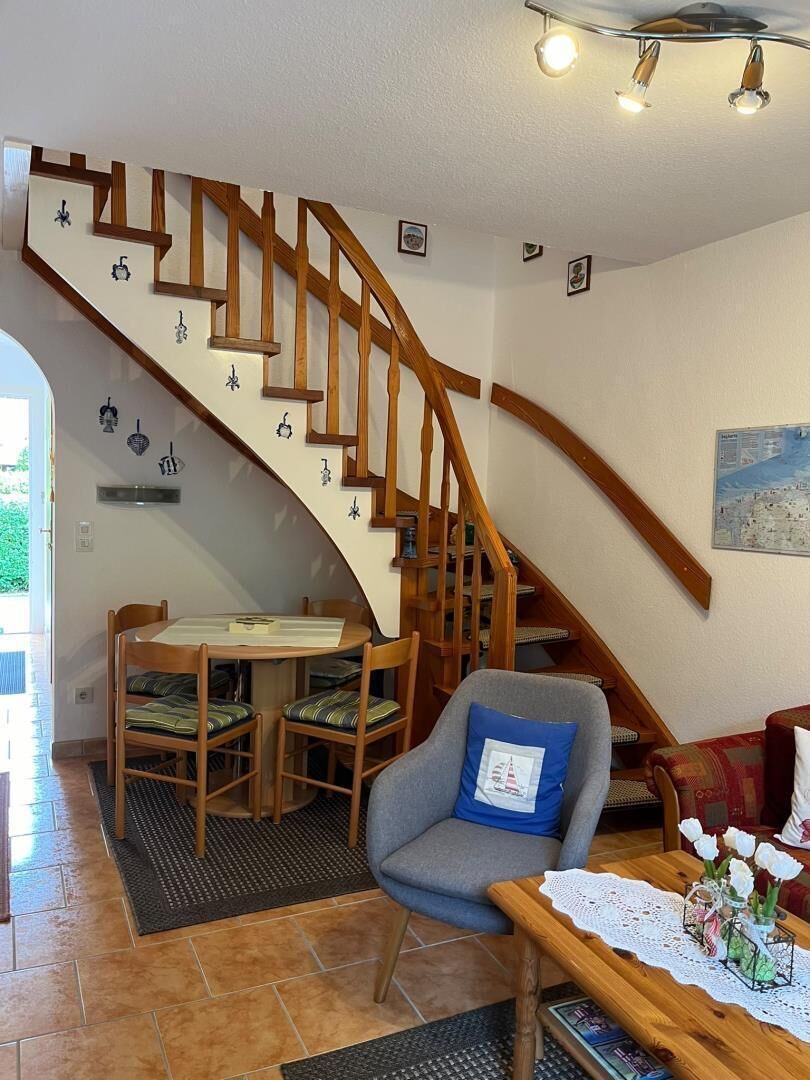 A living room with a wooden staircase, a dining table with chairs, a gray armchair, and a wooden coffee table with a lace table runner.