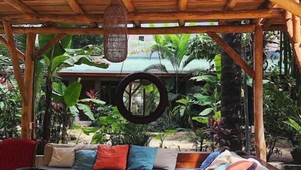 An outdoor seating area with a wooden pergola, cushioned sofas, and a wooden table. - Cabinas Playa Surf Hostel (Cobano)