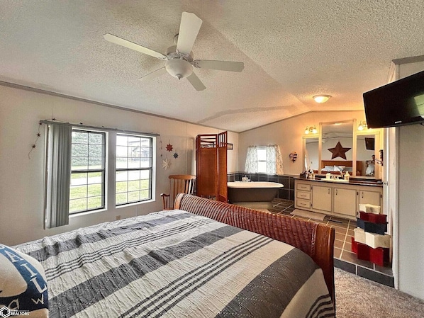 A bedroom with a bed, a ceiling fan, a window, a bathroom with a bathtub, and a television.