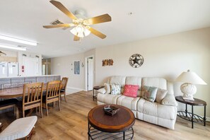 A well-lit living room with a ceiling fan, a dining area with a wooden table and chairs, a sofa with decorative pillows, and a small side table with a lamp. - Coastal Retreat with Pool & Beach Access (Port Aransas)