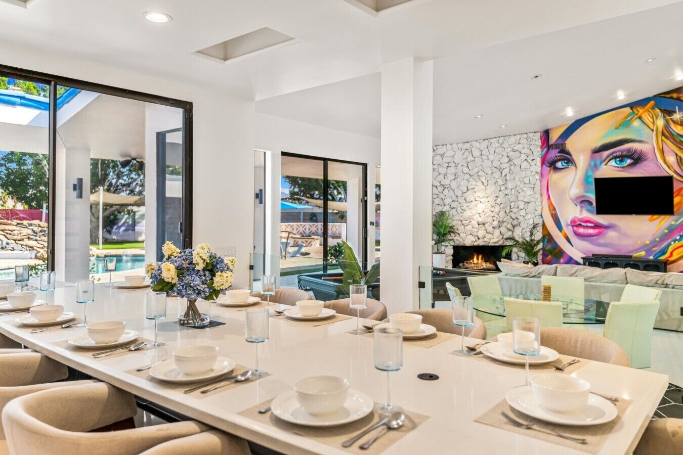 A modern dining area with a long table set for a meal, a fireplace, and a large mural on the wall.