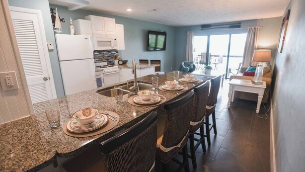 A kitchen with granite countertops, white cabinets, a microwave, and a dining area with a table and chairs. - Steps to Beach, Zen balcony, Pier Park & more (Panama City Beach)