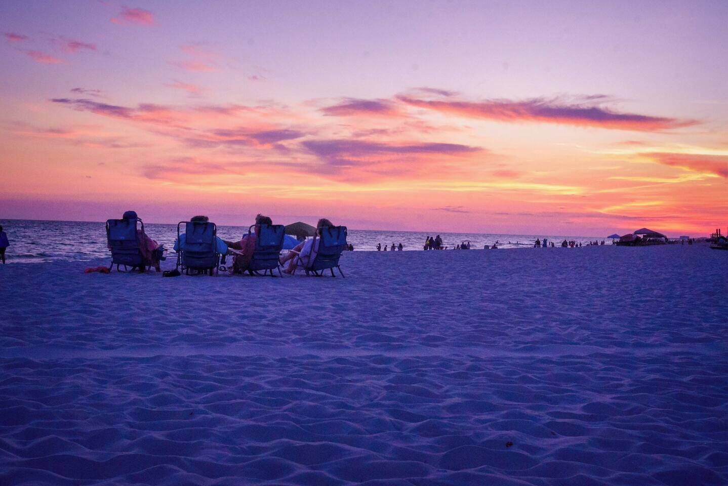A beach scene with people sitting on beach chairs, a few umbrellas, and a sunset in the background.