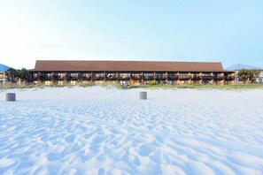Ein Strandhotel mit mehreren Balkonen, einer flachen Dachterrasse und einem sandigen Strand im Vordergrund.