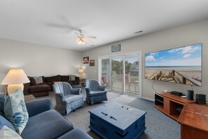 A living room with a ceiling fan, a large TV, and a view of the beach through the sliding glass door. - The Rivalry House 10 Bedroom W/ Private Pool and Short Walk to Beach (Surfside Beach)