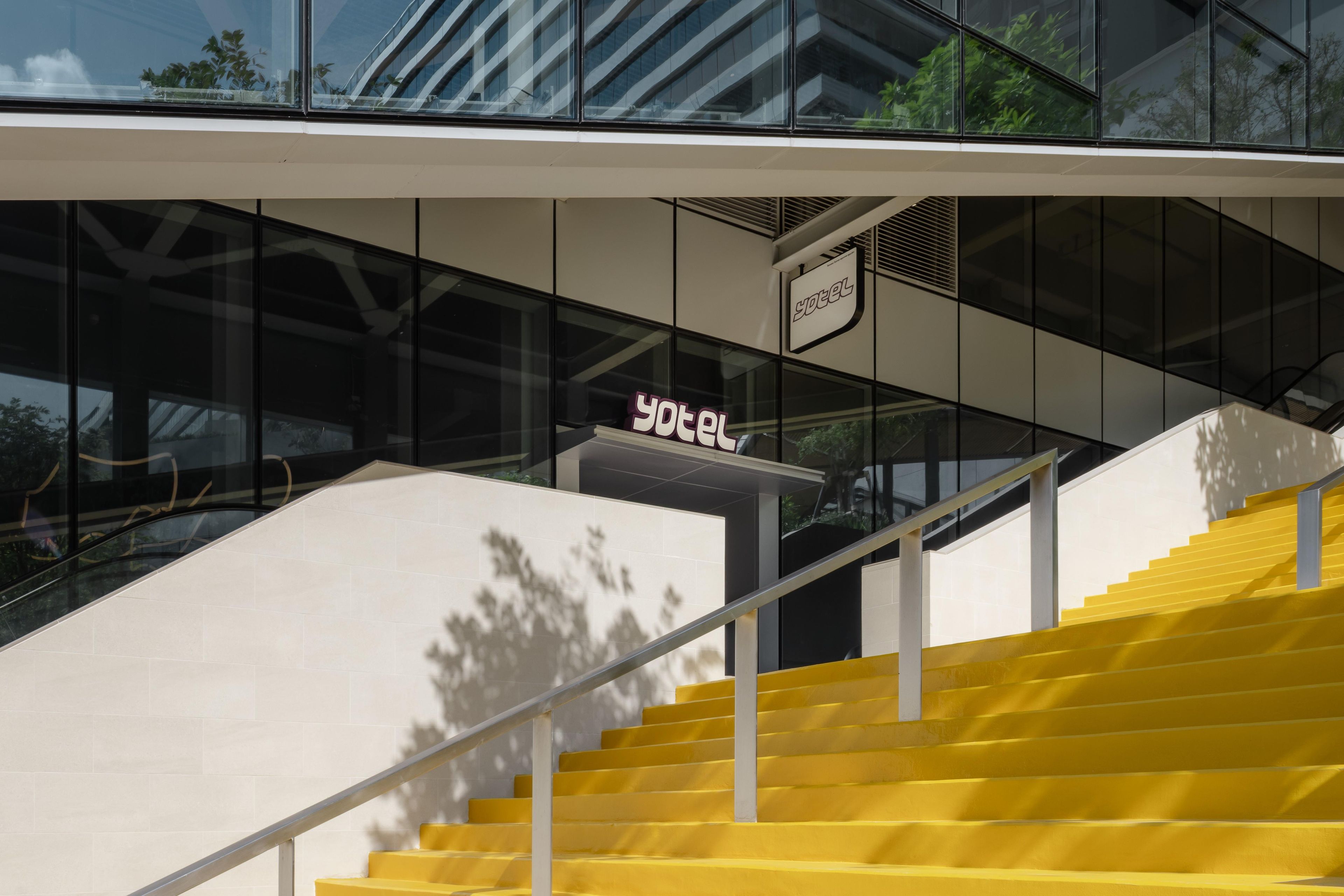 Une entrée d’hôtel moderne avec une façade en verre, une enseigne indiquant qu’il s’agit d’un hôtel, et un escalier avec des marches jaunes.