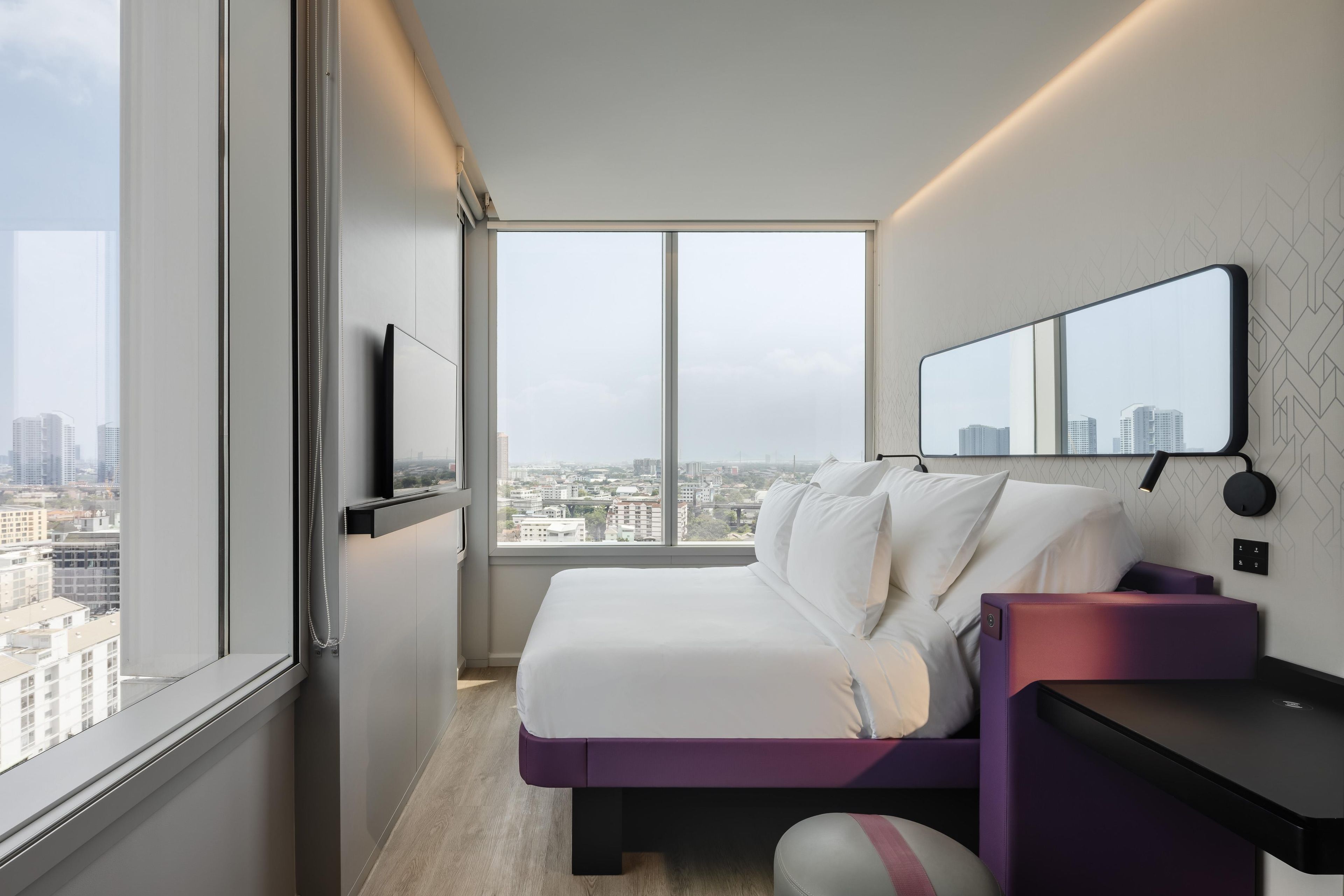 Una habitación de hotel moderna con un ventanal grande, una cama, un escritorio y un espejo.