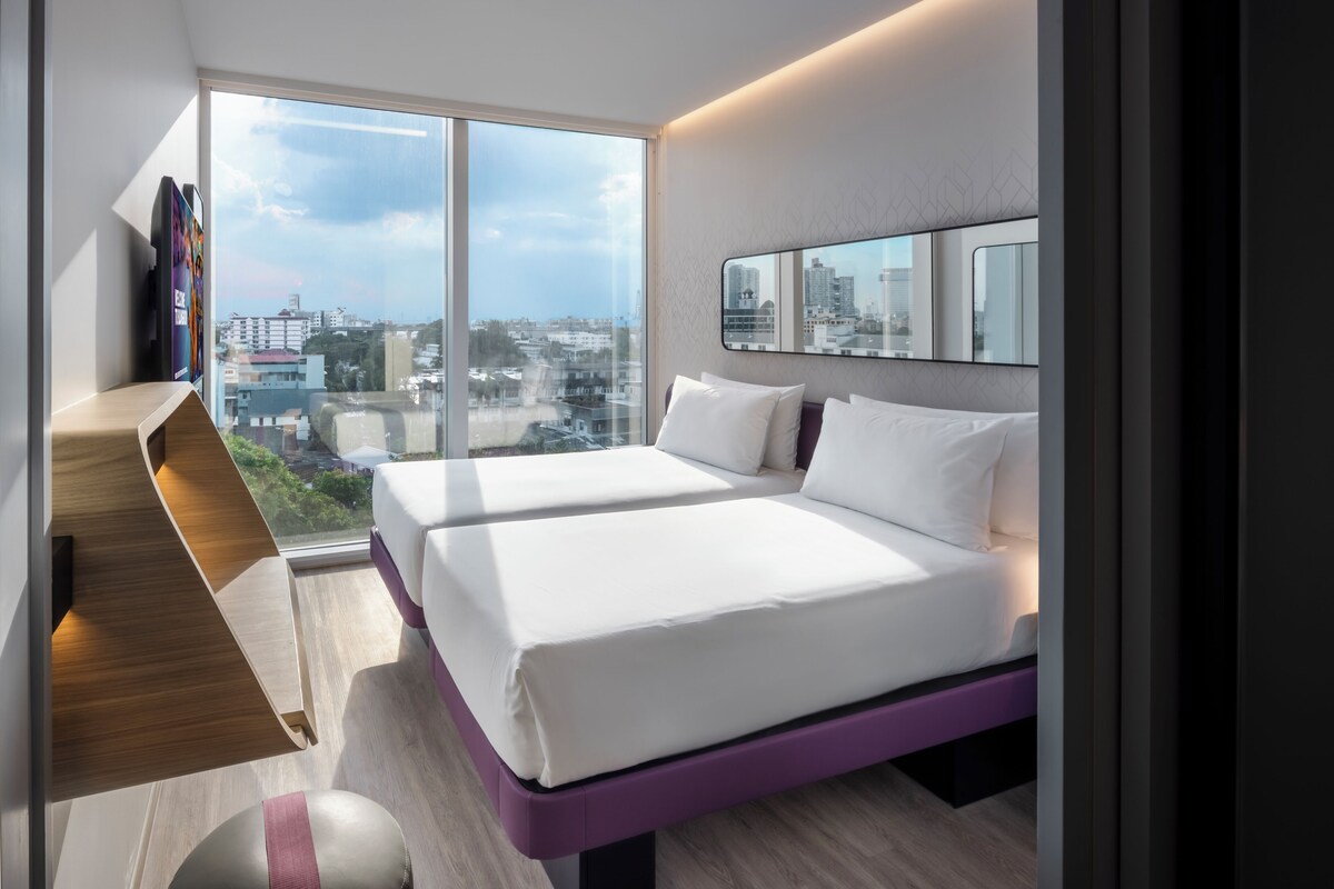 Habitación de hotel moderna con un ventanal grande, dos camas, un banco empotrado y vistas a la ciudad.