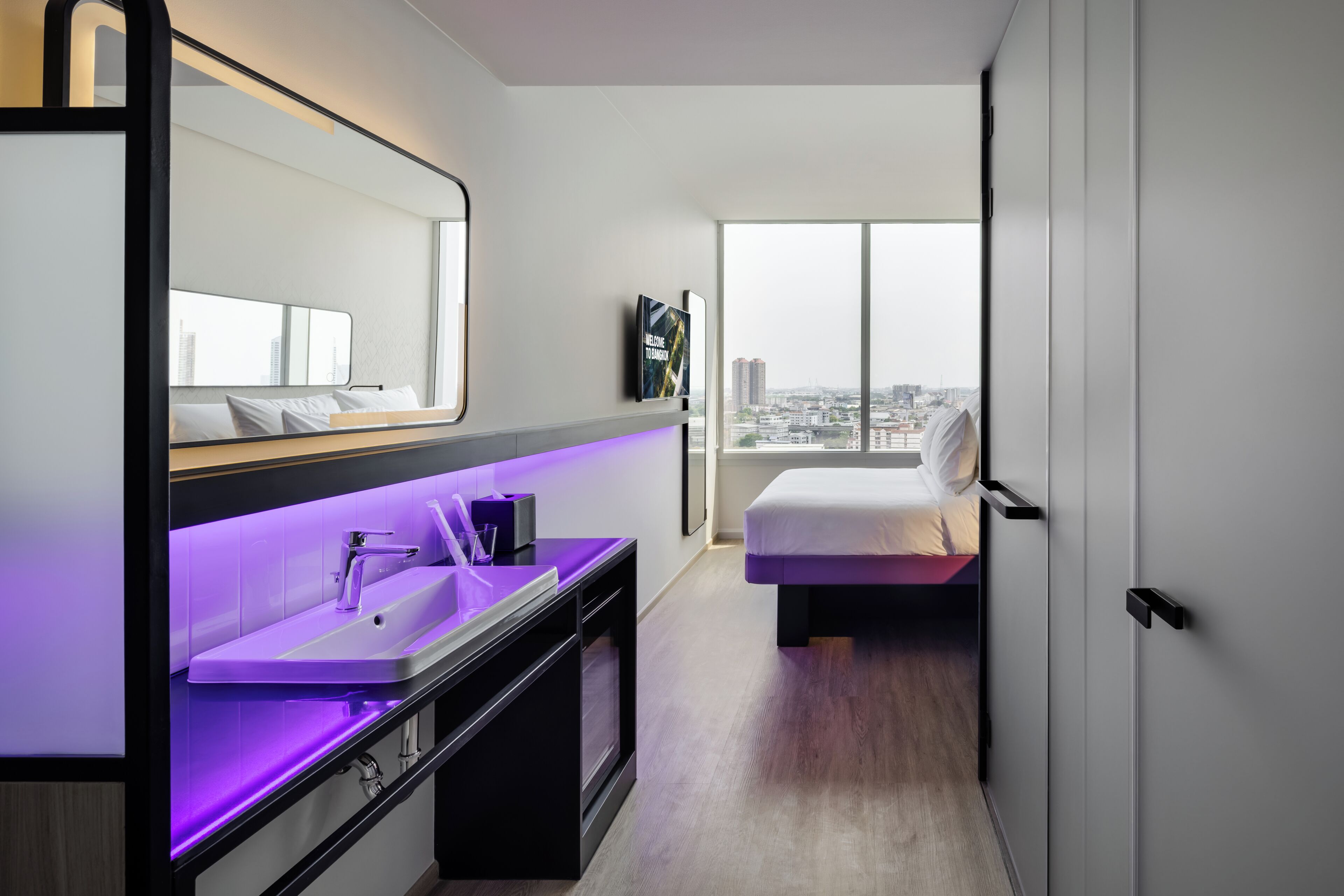A modern hotel room with a large window, a bed, a vanity with a mirror, and a sink.