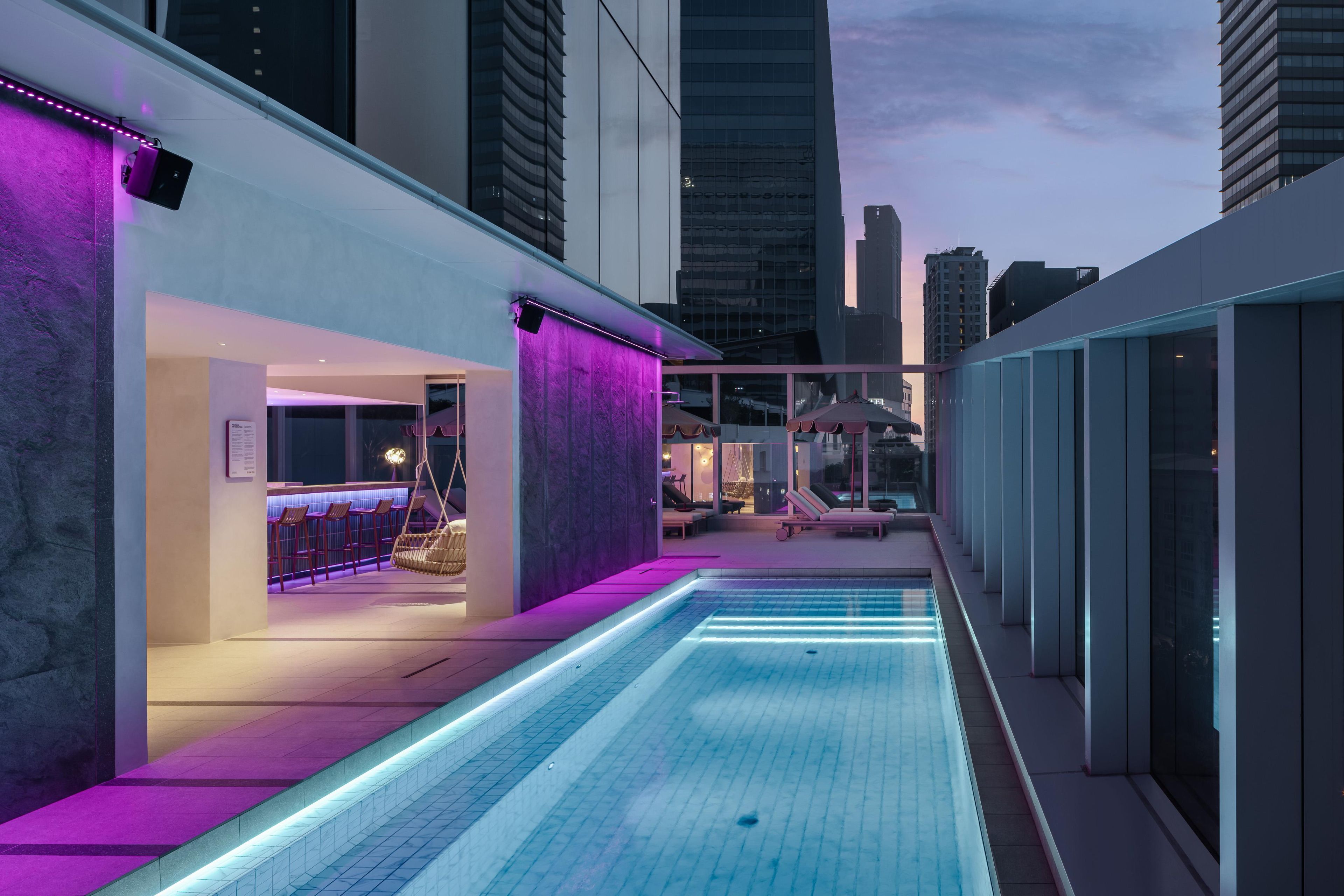 Un espace piscine extérieur moderne, doté d’un bar et de coins salon, entouré d’immeubles de grande hauteur.