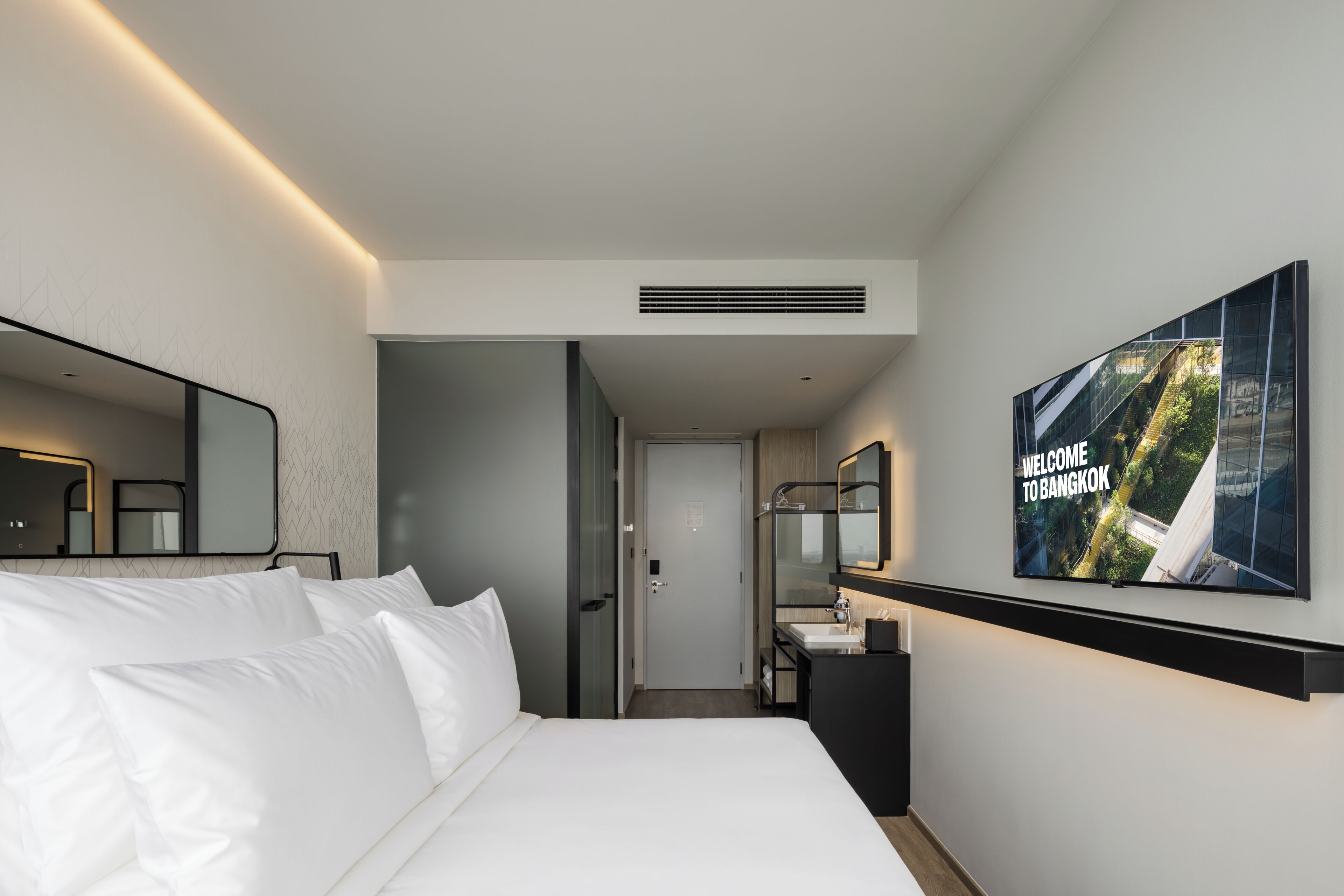 A modern hotel room with a large bed, a bathroom with a mirror and sink, and a wall-mounted TV displaying 'WELCOME TO BANGKOK'.