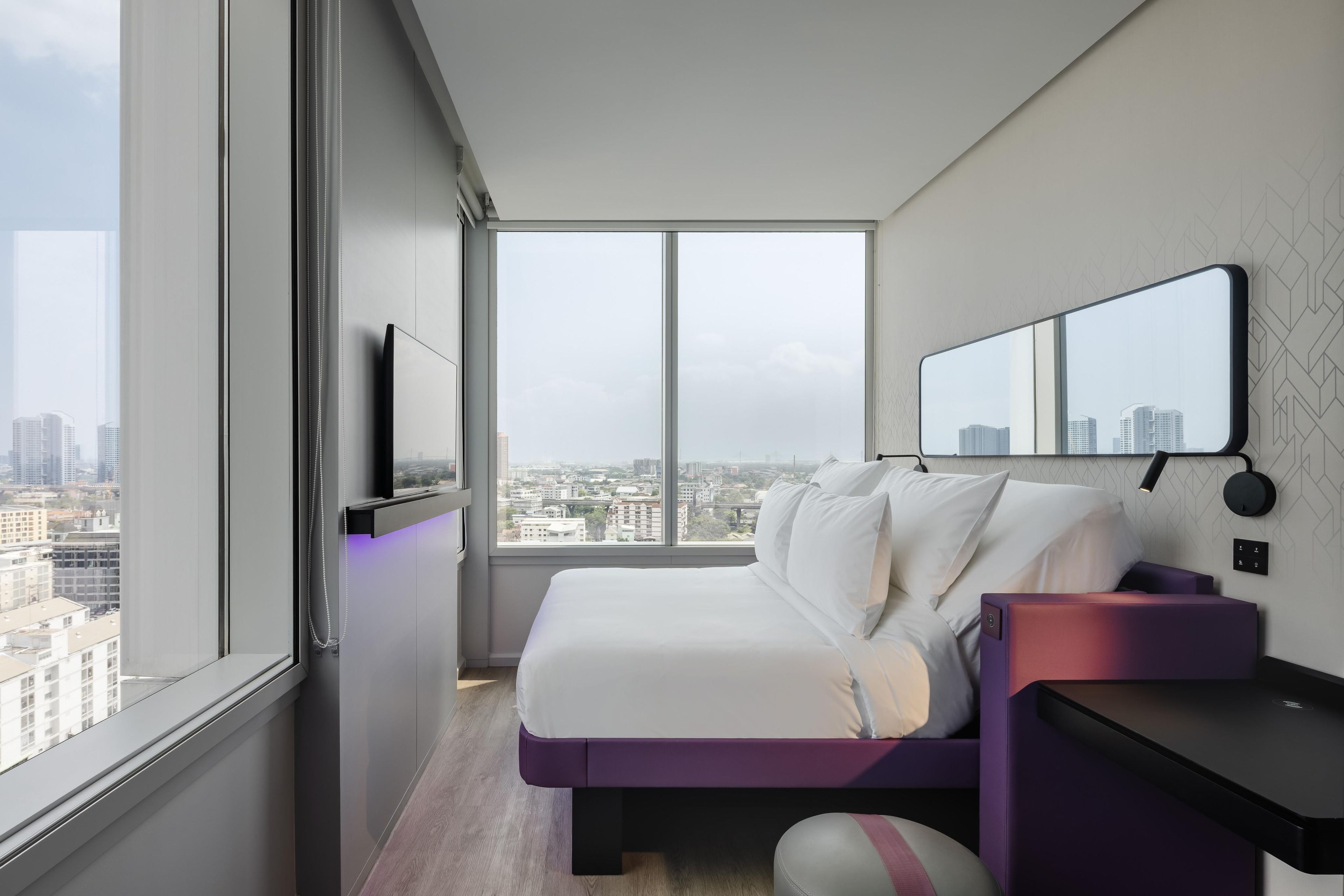 A modern hotel room with a large window, a bed, a desk, and a mirror.