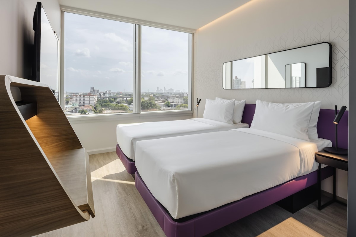 Ein Hotelzimmer mit zwei Betten, einem großen Fenster, einem Schreibtisch und Blick auf die Stadt.