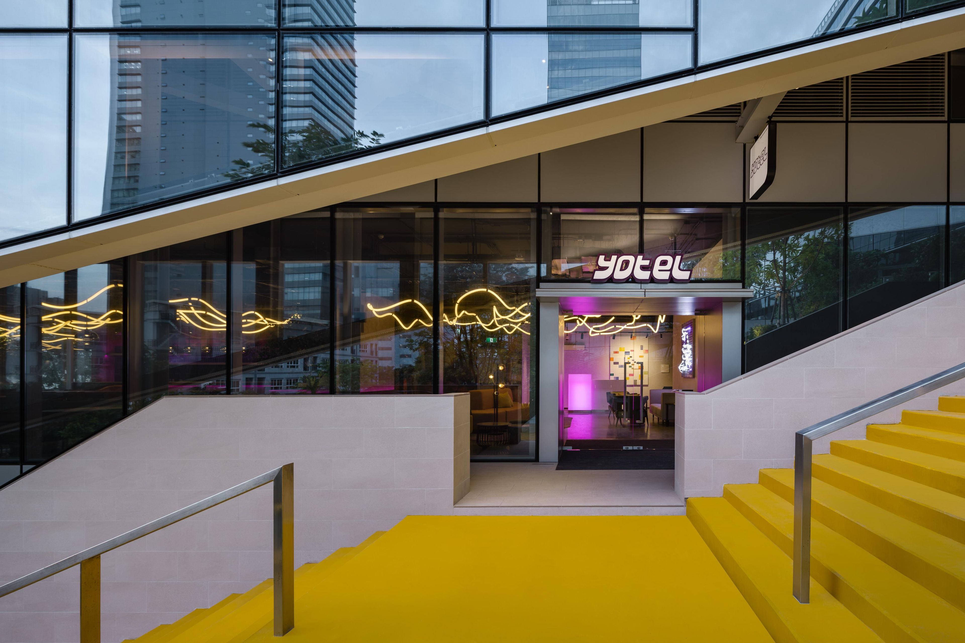 Entrada de hotel com escadas, fachada de vidro e letreiro neon.