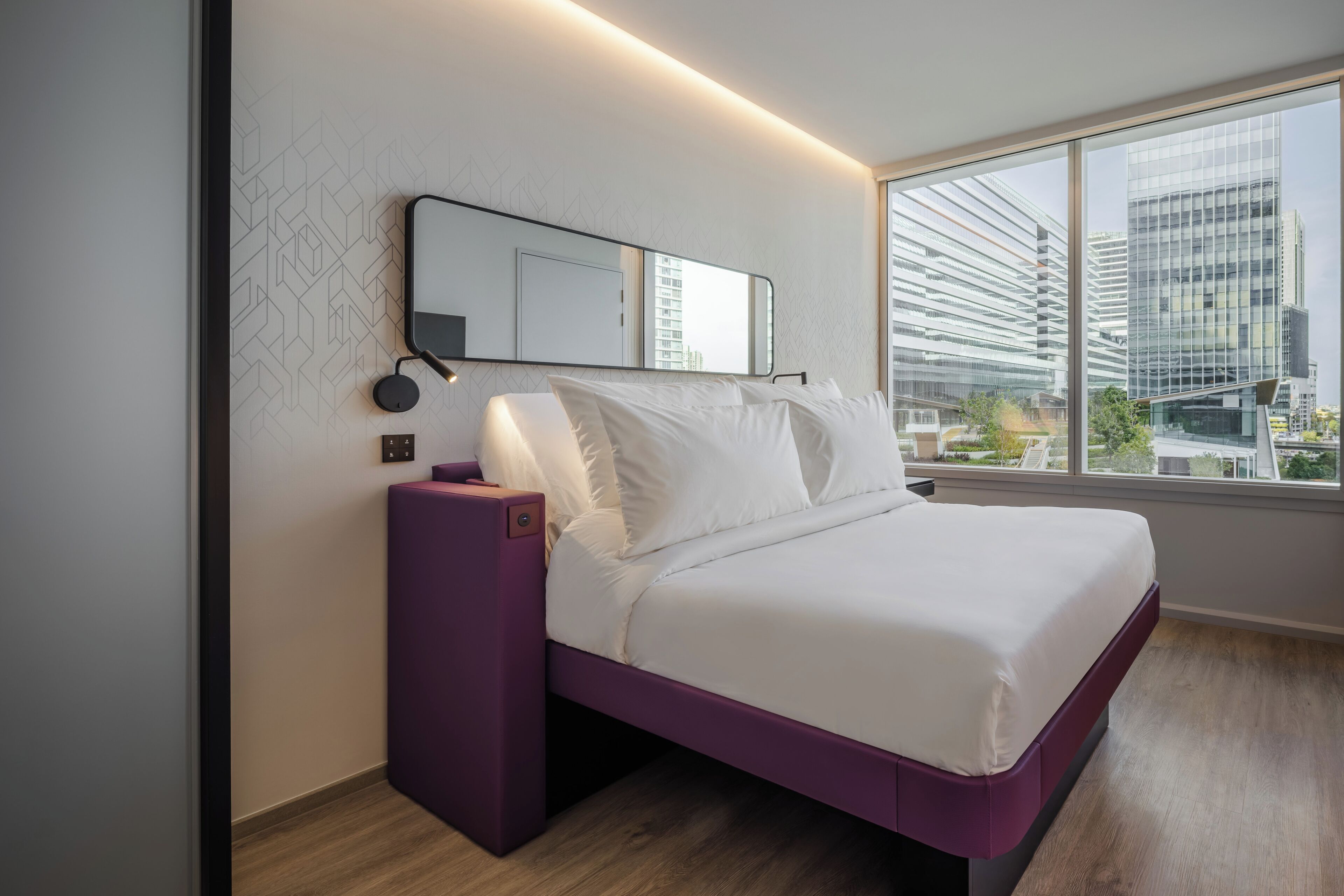 Ein modernes Hotelzimmer mit großem Fenster, einem Bett mit weißer Bettwäsche, einem violetten Nachttisch und einem an der Wand befestigten Spiegel.