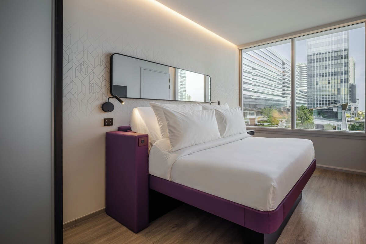 Una habitación de hotel moderna con un ventanal grande, una cama con ropa de cama blanca, una mesita de noche morada y un espejo montado en la pared.