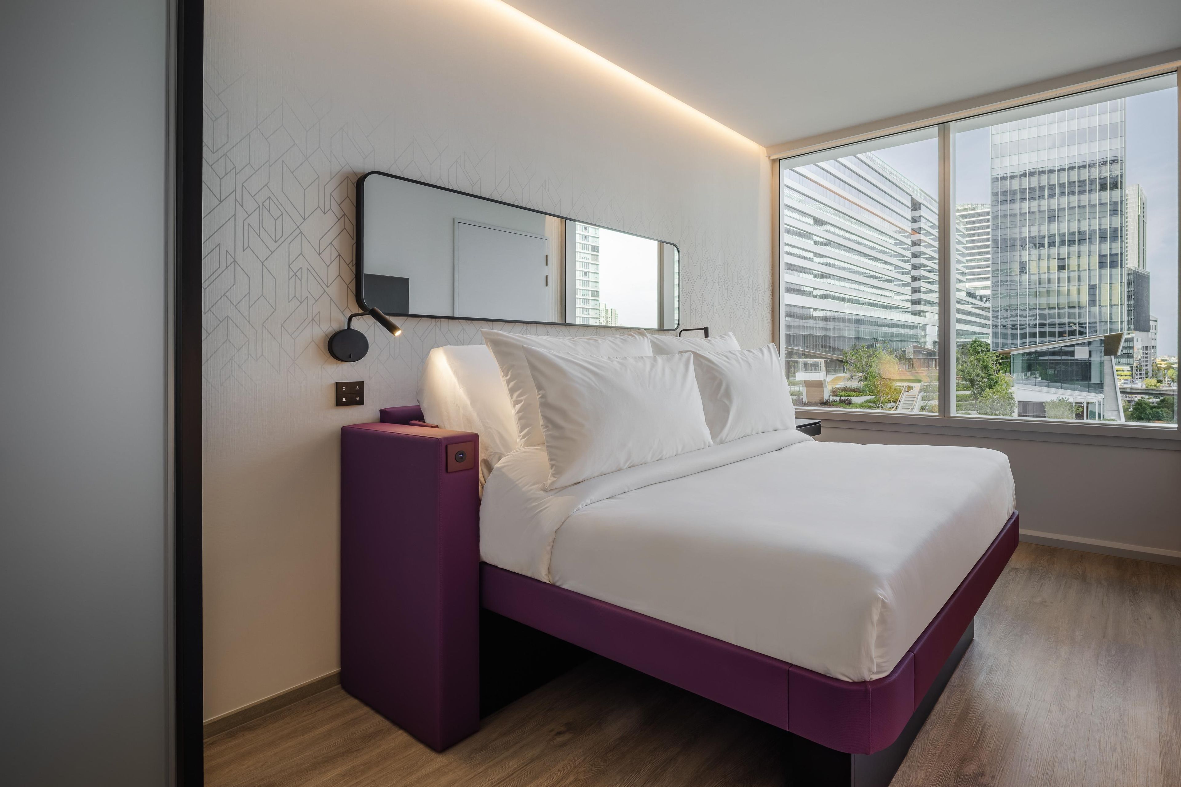 A modern hotel room with a large window, a bed with white bedding, a purple nightstand, and a wall-mounted mirror.