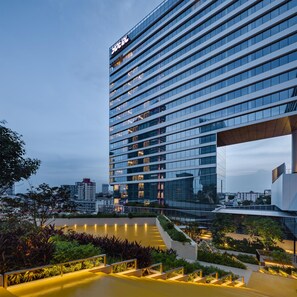 A modern hotel building with a prominent 'HOTEL' sign at the top, multiple floors, and a landscaped outdoor area with pathways and greenery.