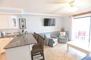 A kitchen with a granite countertop, a dining area with a blue sofa, and a TV mounted on the wall. - Beachfront Condo, Gulf View, Balcony, Beach Chairs, Heated Pool, Jacuzzis (Panama City)