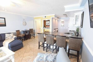 A living room with a sofa, chairs, a dining table, and a kitchen area with a sink and appliances. - Beachfront Condo, Gulf View, Balcony, Beach Chairs, Heated Pool, Jacuzzis (Panama City)