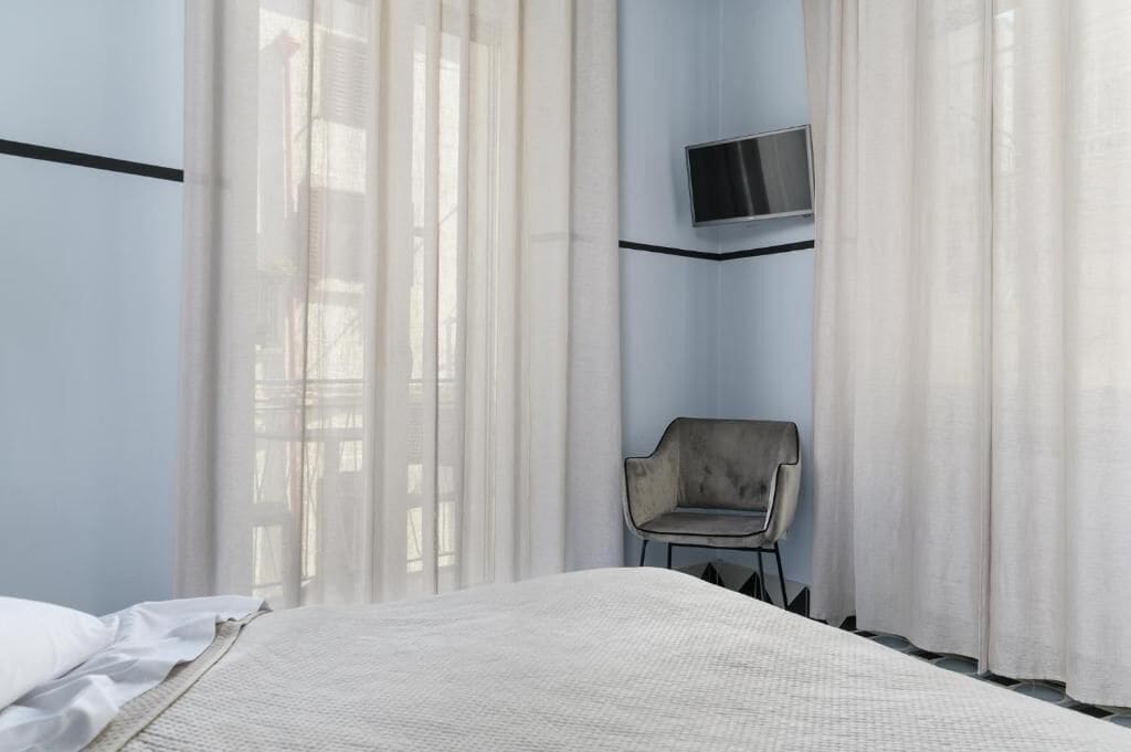 Ein Hotelzimmer mit Bett, einem Sessel, einem an der Wand befestigten Fernseher und durchscheinenden Vorhängen.