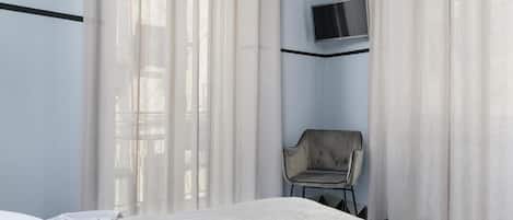 A hotel room with a bed, a chair, a TV mounted on the wall, and sheer curtains.