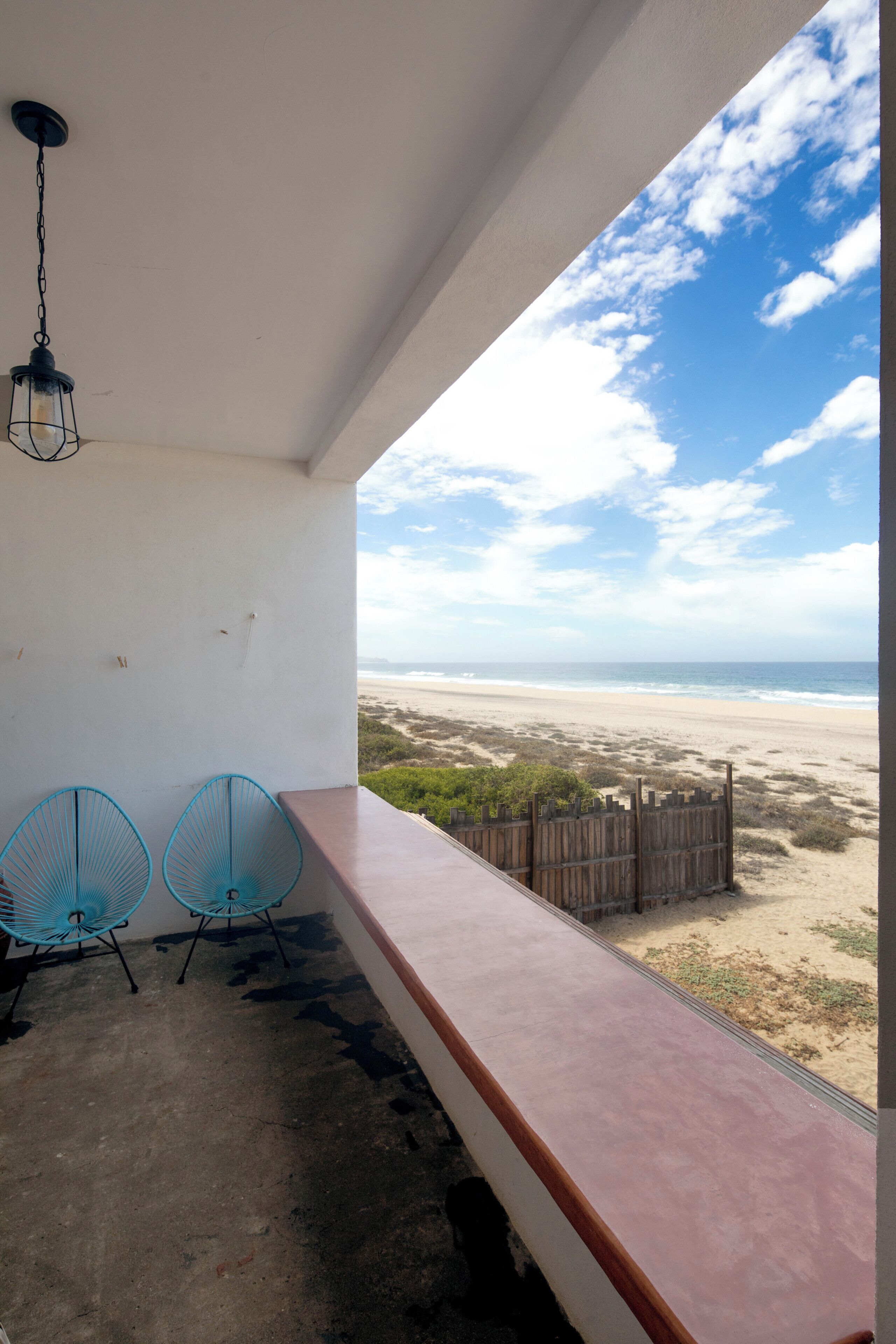 A balcony with a view of the beach, two blue chairs, and a hanging light fixture.