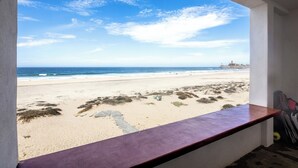 Blick auf den Strand von einem Zimmer aus, mit einem Fenster zum Meer, einer Holzbank und einem Surfbrett, das an der Wand lehnt.