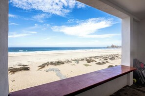 Blick auf den Strand von einem Zimmer aus, mit einem Fenster zum Meer, einer Holzbank und einem Surfbrett, das an der Wand lehnt.