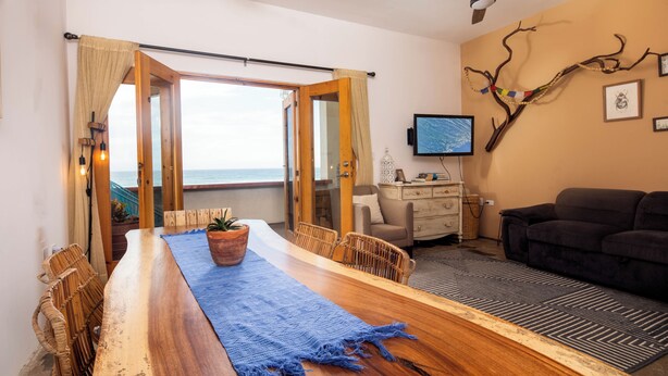 Ein Zimmer mit Esstisch aus Holz, Korbstühlen, einer Couch, einem Fernseher und Meerblick.
