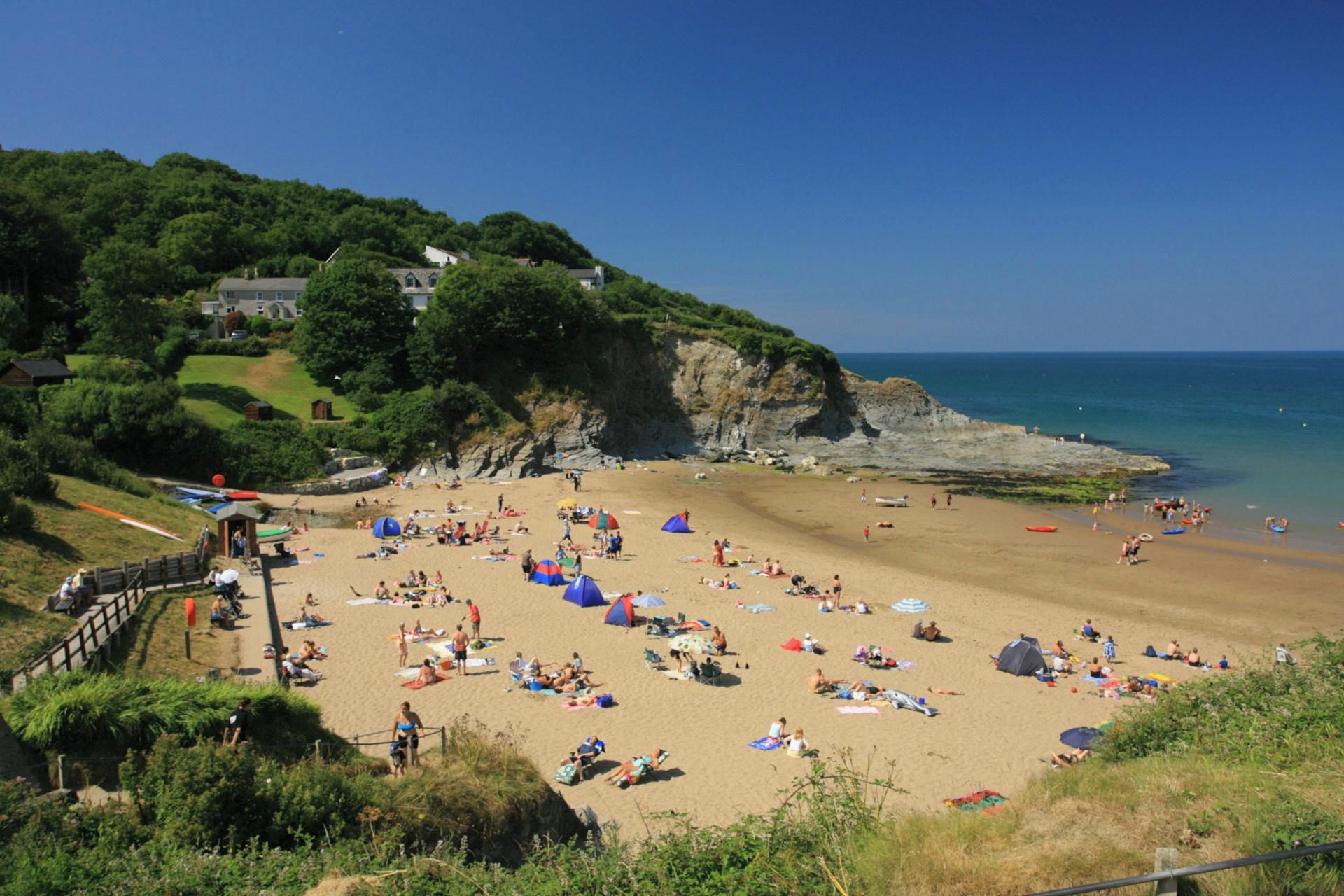A beach with people sunbathing and swimming, a rocky cliff, and a clear sky.