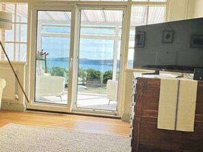 A room with a television on a wooden cabinet, a sliding glass door leading to a balcony with a view of the sea, and a white chair on the balcony. - 3 Hilltop Way, Aberporth (Aberporth)