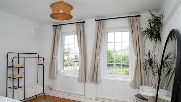A room with a bed, a radiator, a window with curtains, a hanging light fixture, and a plant. - 3 Hilltop Way, Aberporth (Aberporth)