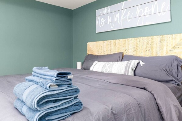 A neatly made bed with a grey comforter and pillows, a stack of folded towels, and a wall with a sign that says 'shhh... no wake bone.' - Lakin` It Easy | Fire Pit+H20 Trampoline+GameRoom (Henrico)