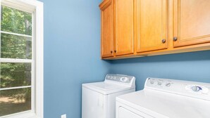 A laundry room with two white washers and dryers, wooden cabinets, and a window with a view of trees.