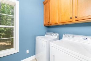A laundry room with two white washers and dryers, wooden cabinets, and a window with a view of trees.