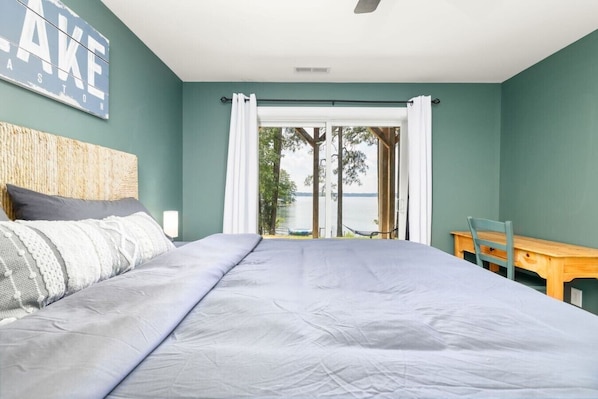 A bedroom with a bed, a wooden chair, a table, a view of the outdoors, and a sign on the wall. - Lakin` It Easy | Fire Pit+H20 Trampoline+GameRoom (Henrico)