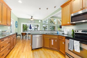 A kitchen with wooden cabinets, a stainless steel dishwasher, a stove, and a large window overlooking a green outdoor area.