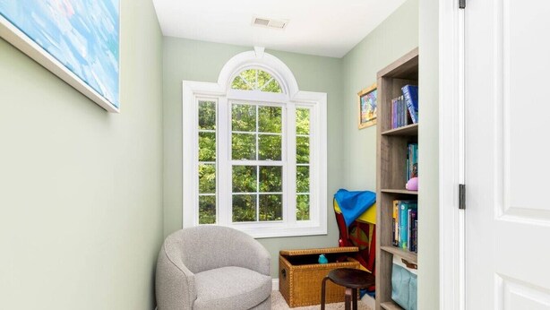 A room with a large window, a grey armchair, a small table, a bookshelf, and a door.