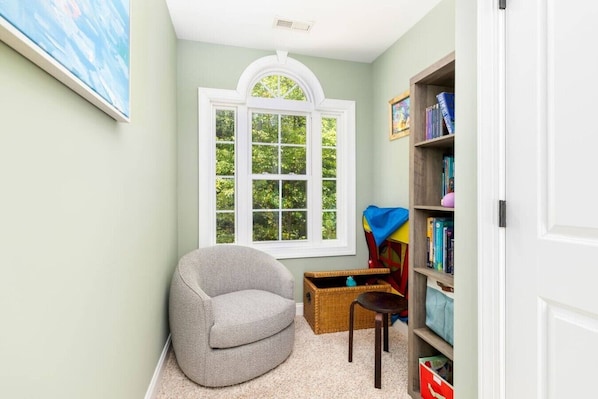 A room with a large window, a grey armchair, a small table, a bookshelf, and a door.