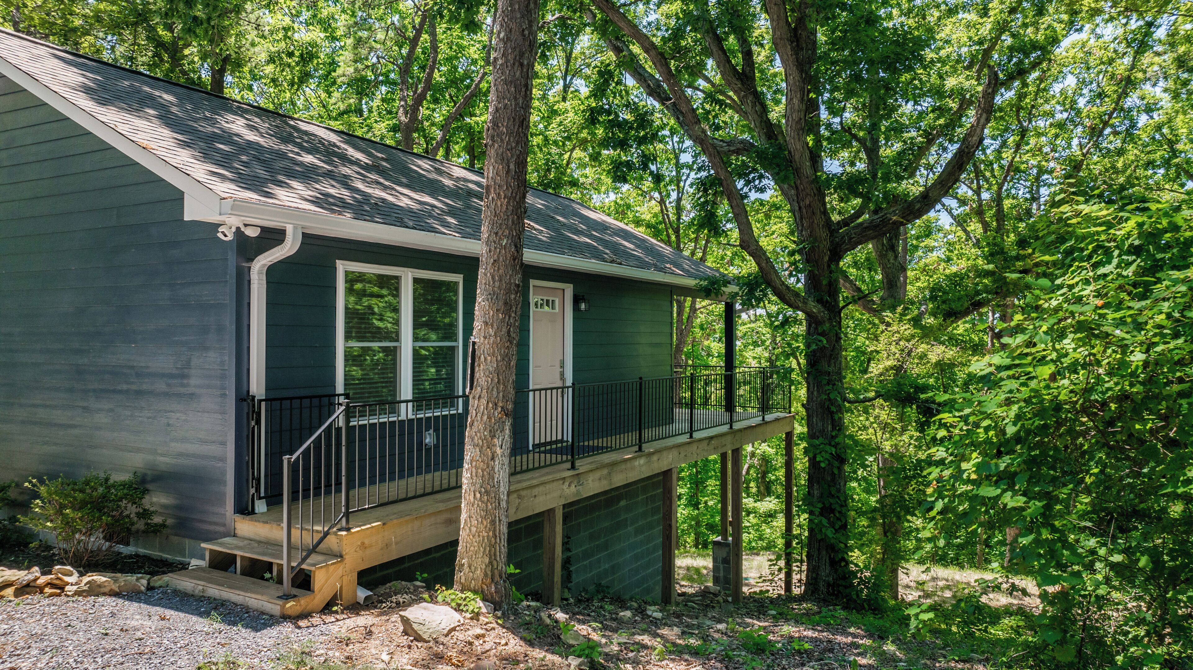 A small, single-story cabin with a front porch and a deck, surrounded by dense forest.