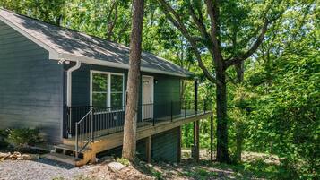 A small, single-story cabin with a front porch and a deck, surrounded by dense forest.